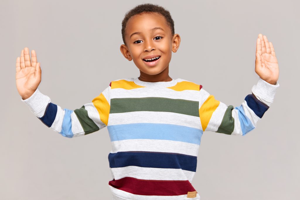 Body language. Studio shot of handsome adorable Afro American boy in striped pullover keeping hands wide apart as if holding something very large in size, measuring, having excited expression